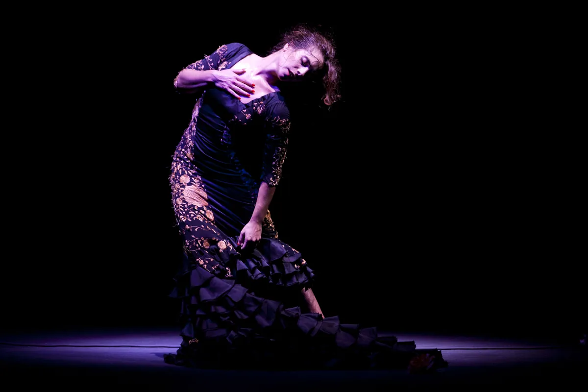 Tanečnice flamenca v tmavých vyšívaných šatech se ohýbá v pase s jednou rukou na hrudi, sama ve světle reflektoru na černém jevišti
