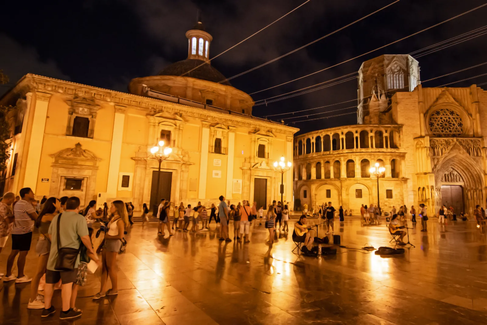 Plaza de la Virgen i Valencia om natten, med en menneskemængde på den lampelyste plads foran den kuplede basilika og katedralens gotiske portal