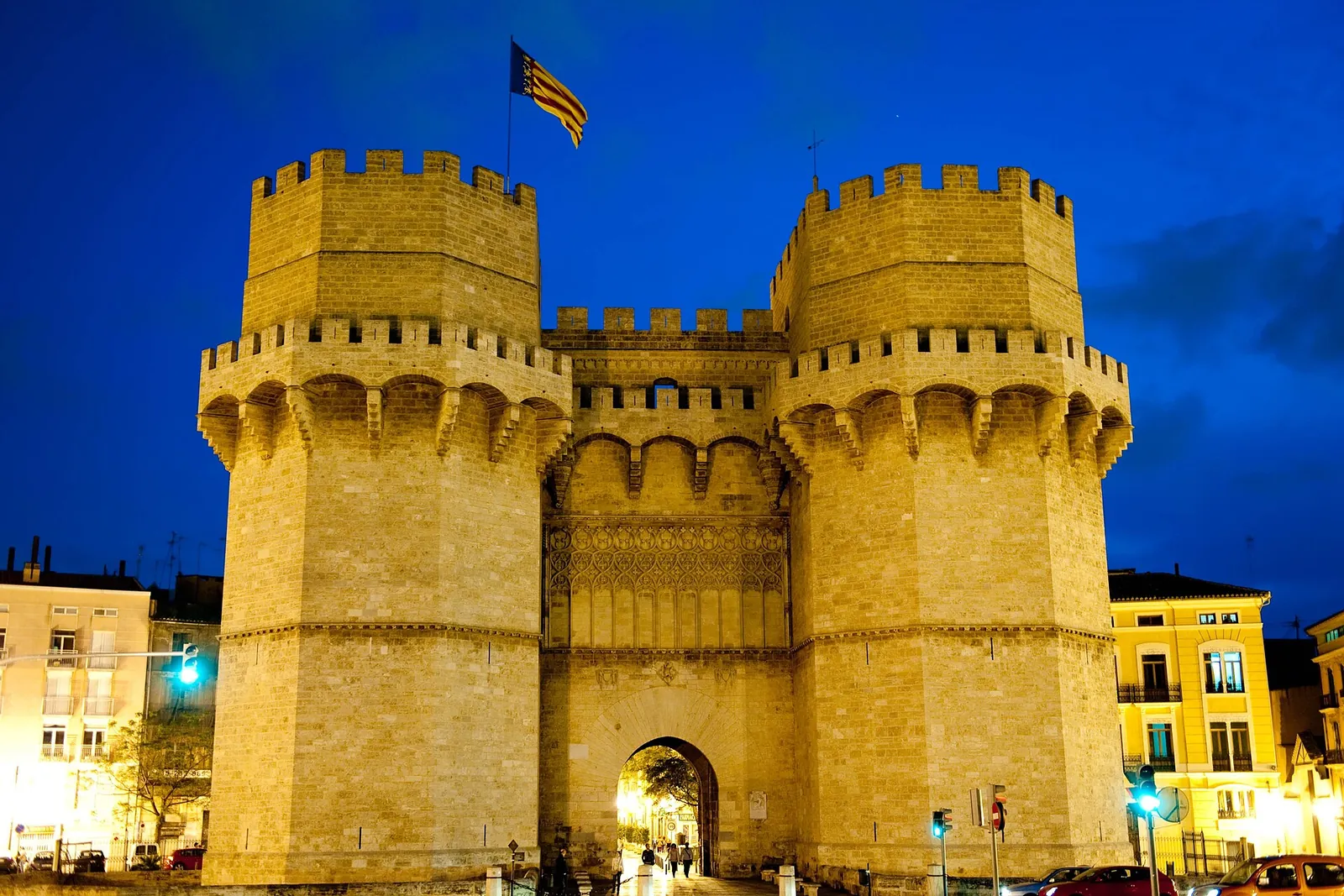 Torres de Serranos i tusmørket med et valenciansk flag på venstre tårn og gult oplyst sten under en dybblå himmel
